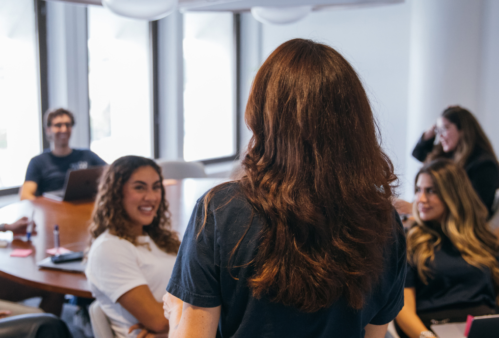 A group of people sitting around a table and a woman wearing a black t-shirt is addressing something to them.