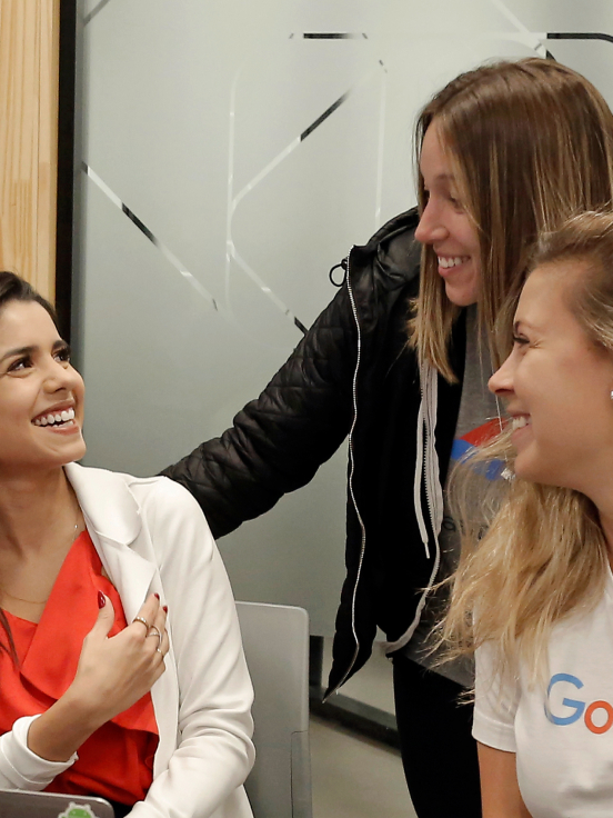 Três mulheres sentadas em uma sala de reuniões conversando e sorrindo.