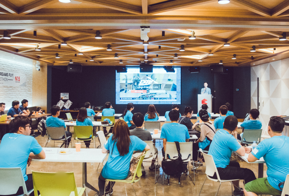 A group of people wearing same sky blue t-shirt listening to a man presenting something