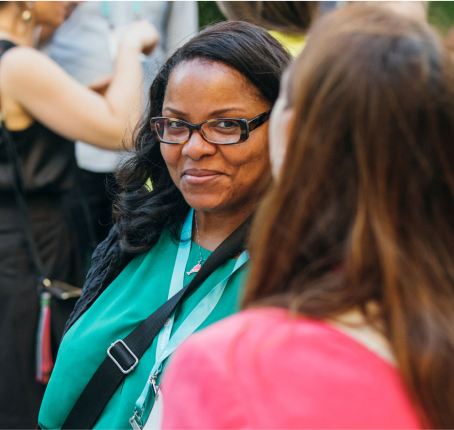 A dark woman with long hair and wearing spectacles smiling at other woman in crowd.