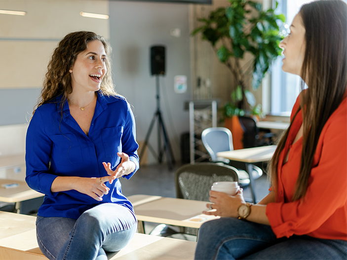 A woman wearing blue top in focus talking to another woman facing away from the camera.