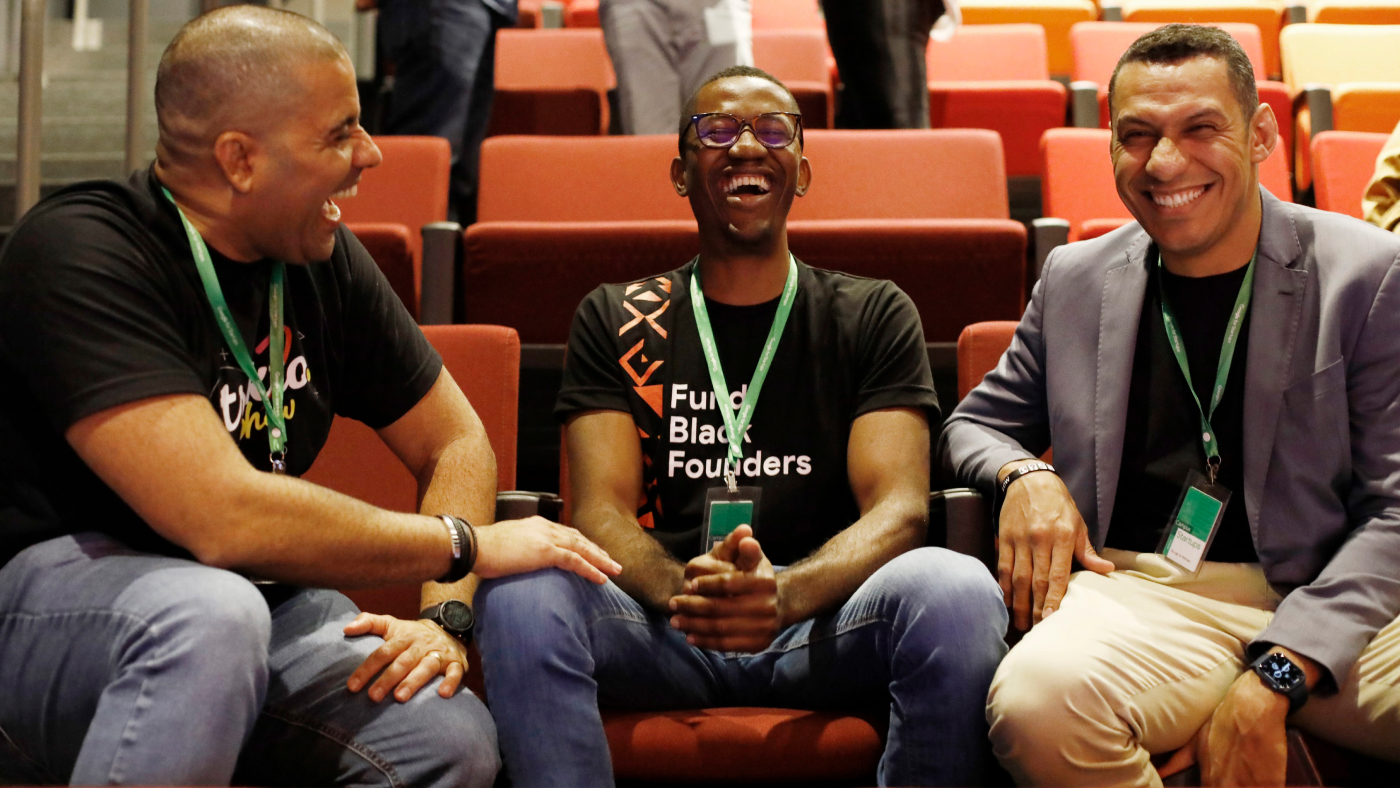 Three men discussing and laughing while sited in conference room, and one man looking into the camera,.