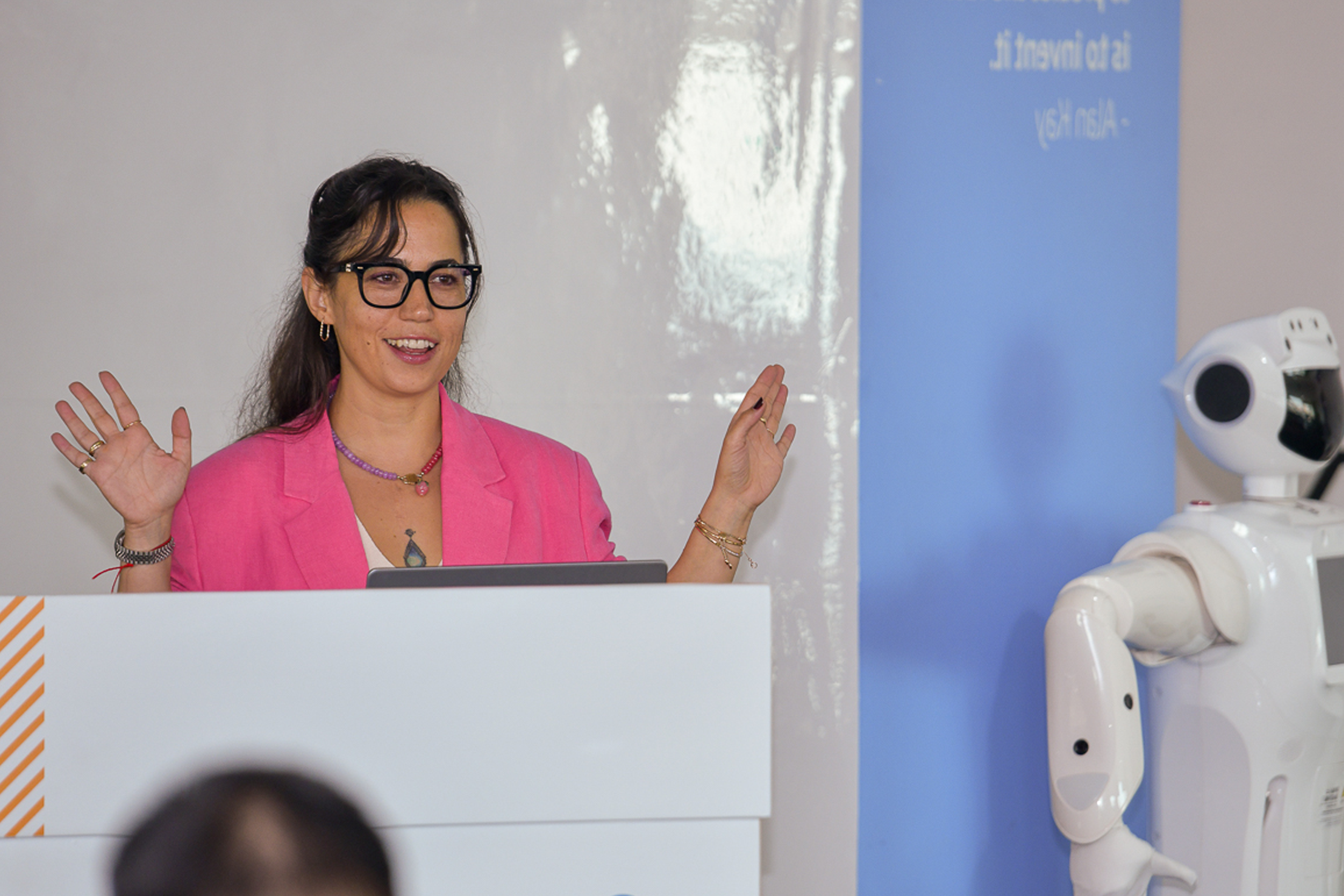 The female presenter, dressed in a pink suit, is leading the event and delivering her segment.