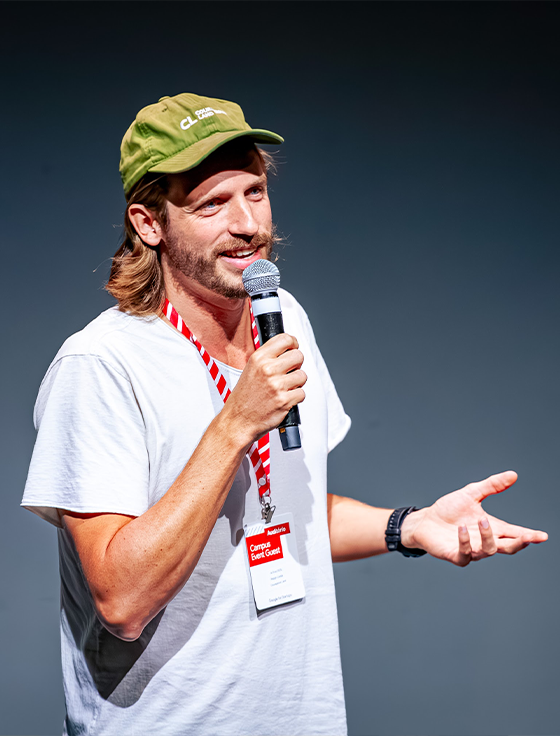 Um apresentador se dirige à plateia do palco, usando um boné.