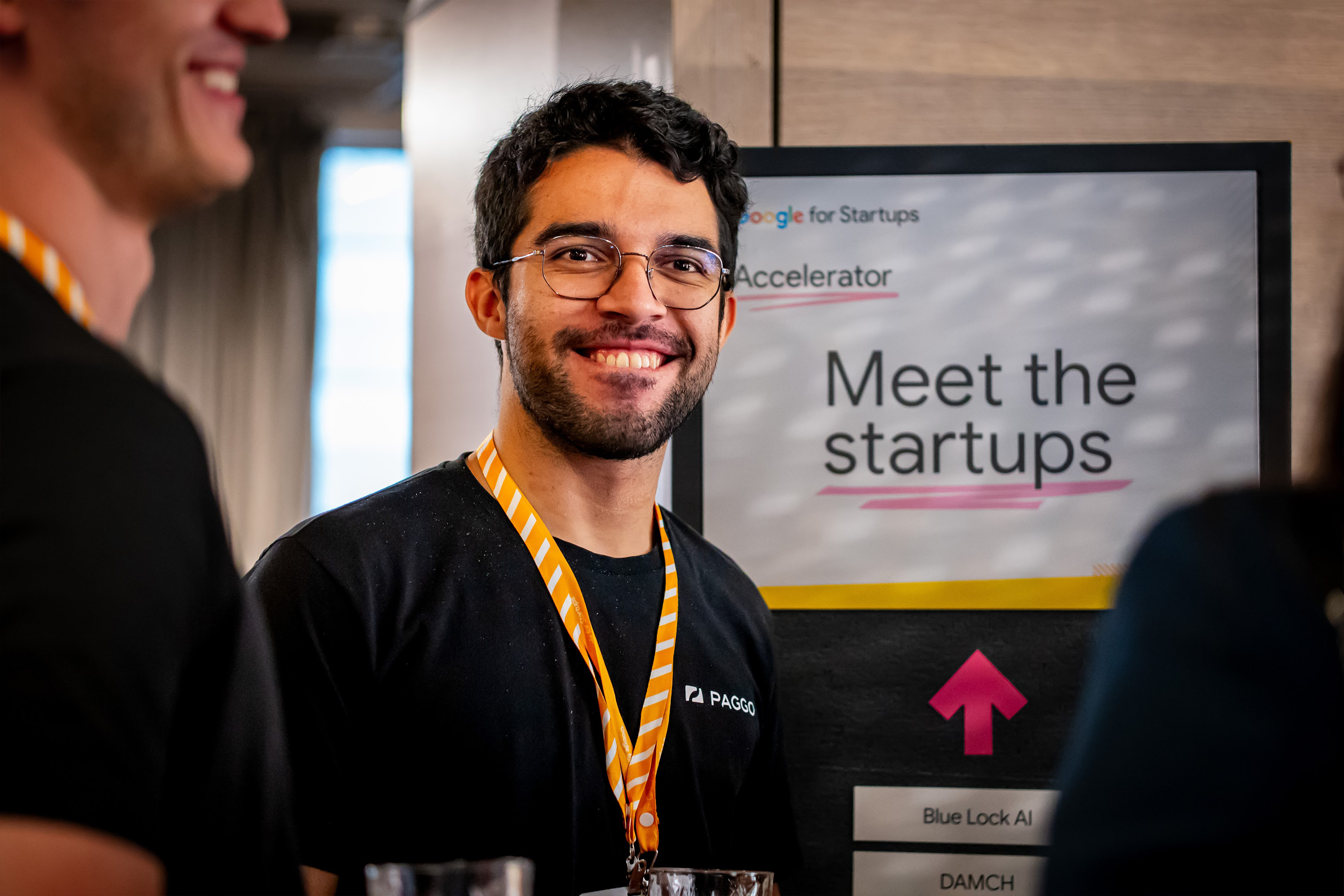 Um participante do evento está em pé ao lado de uma grande tela que exibe a mensagem "Conheça as startups".