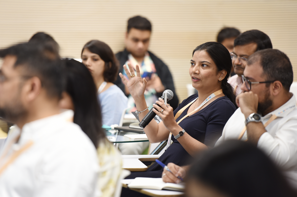 A woman speaking in mic surrounded by other men and women while attending some session