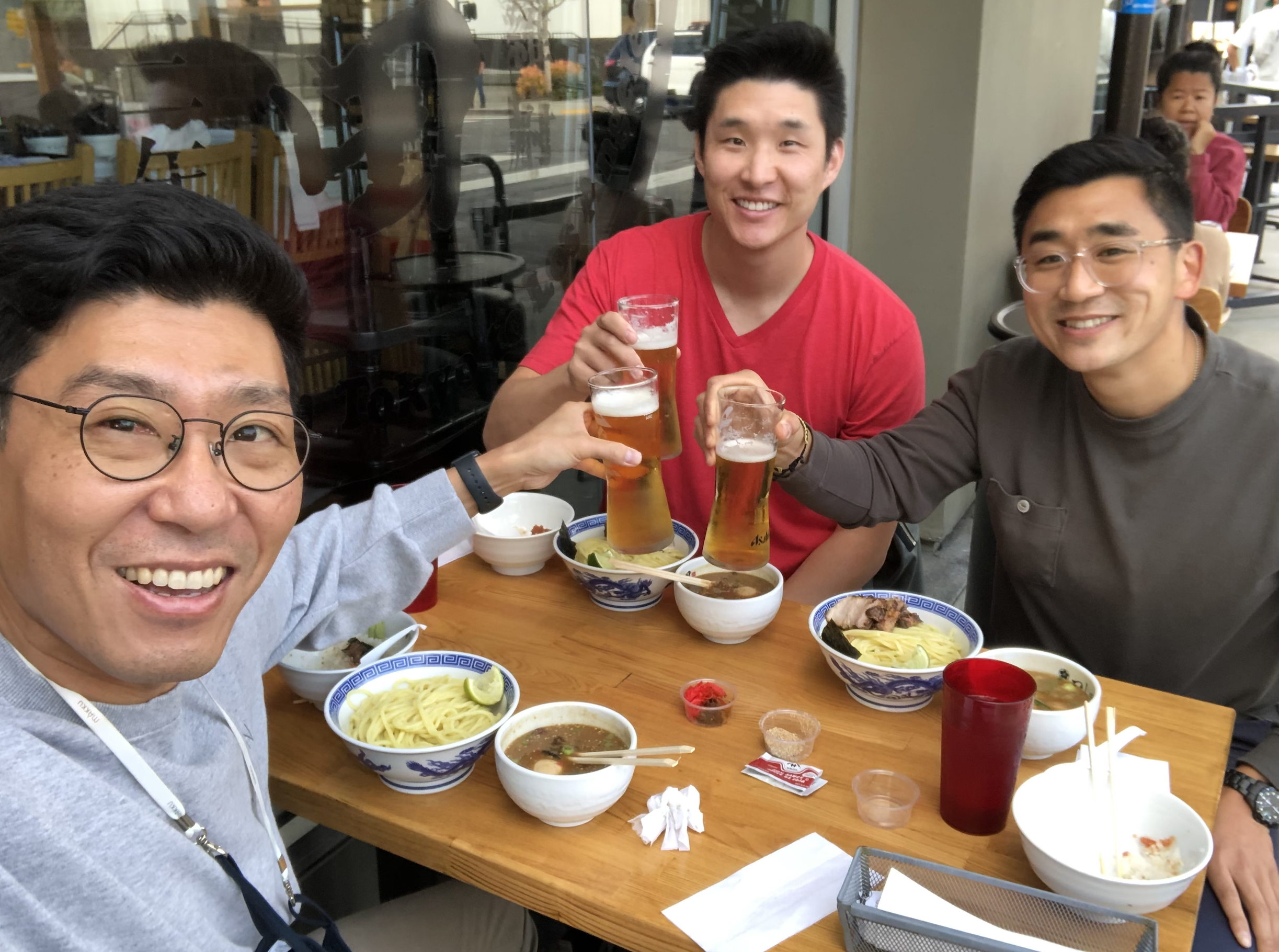 Three men smile looking at the camera while raising their beers together over a table with bowls of noodles.