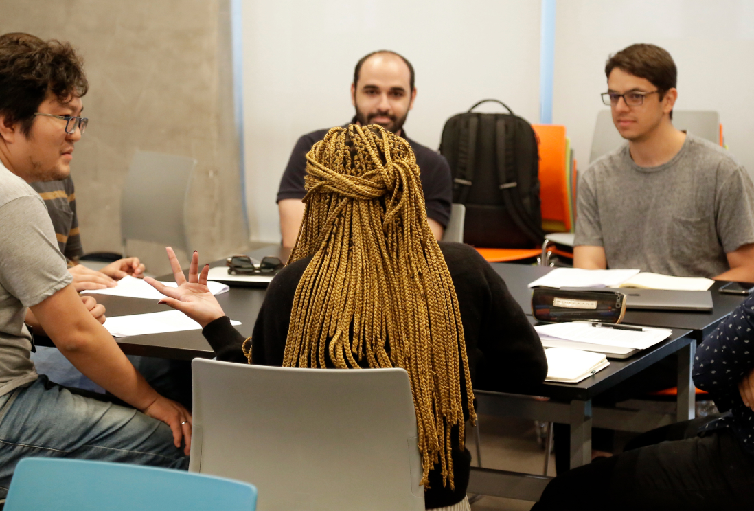 A woman sitting in the center around a group back to the camera is saying something, and other mans looking into her.